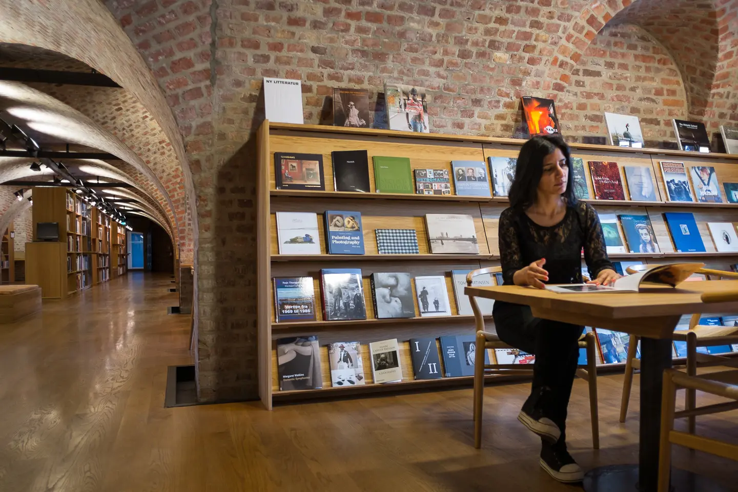 En kvinne sitter ved et bord i et bibliotek og leser en bok. Fotografi.