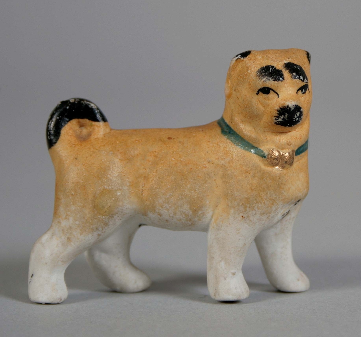 Stående hund med högervridet huvud i oglaserat porslin. Målad gulbrun med svans och ansiktsdetaljer i svart. Halsband i grönt med guldrosett.