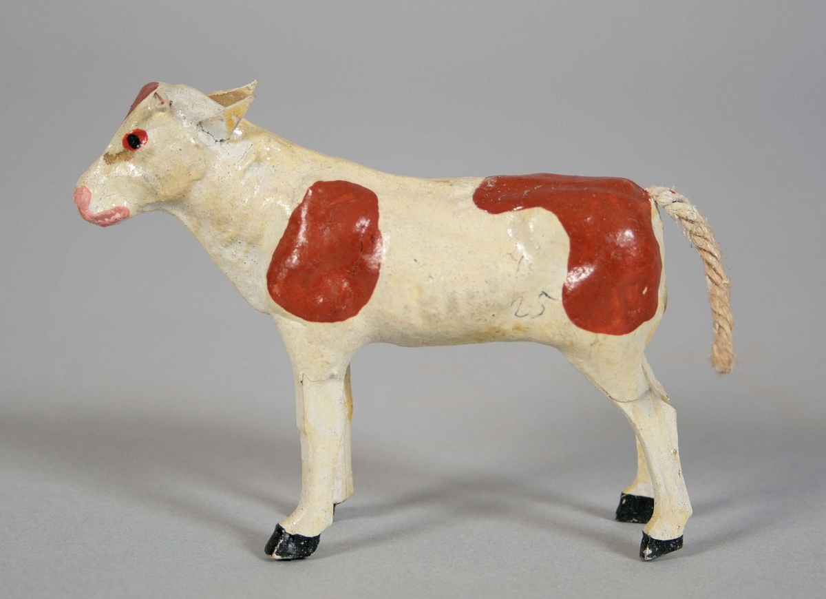 Stående ko i papiermaché med svans av ofärgad hampa. Målad vit med fläckar i rödbrunt. Höger horn avbrutet.