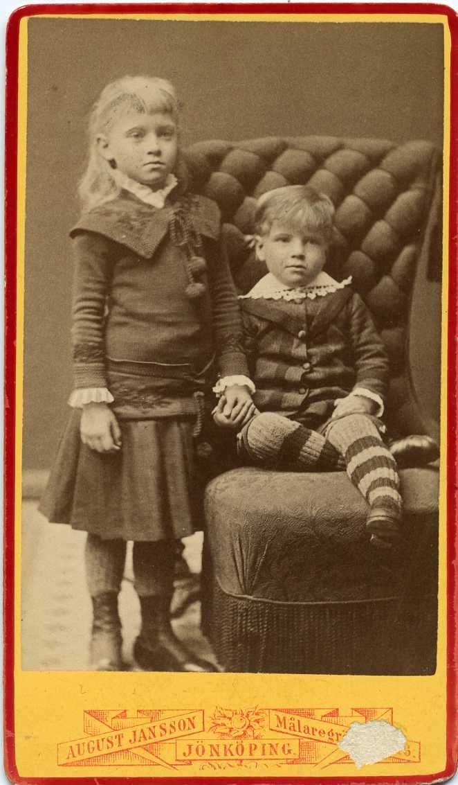 En liten flicka står intill en mindre pojke som sitter i en emma, de håller varandra i handen. De är syskon