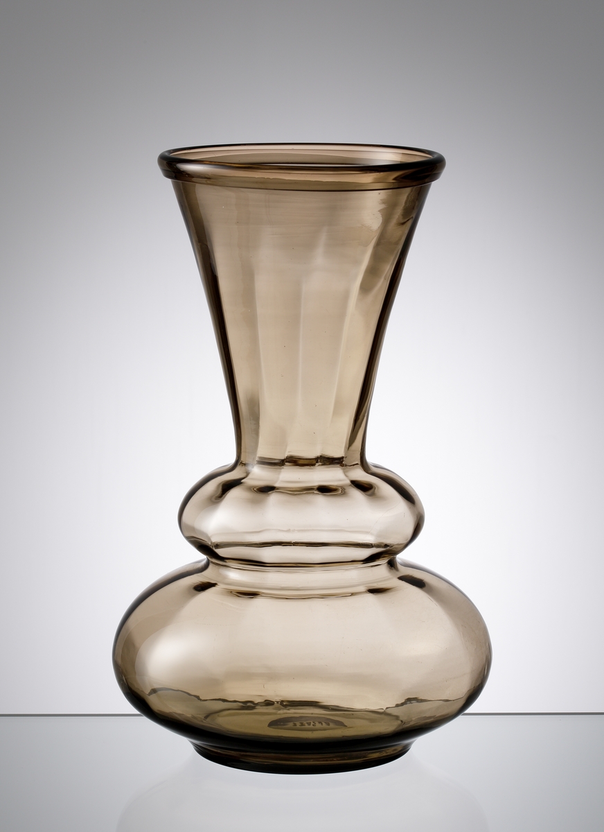 Formgiven av Edward Hald. Blomglas i bruntonat sodaglas. Trumpetformad hals med omvikt mynningskant.
I nedre delen två vulster. Optikblåst.