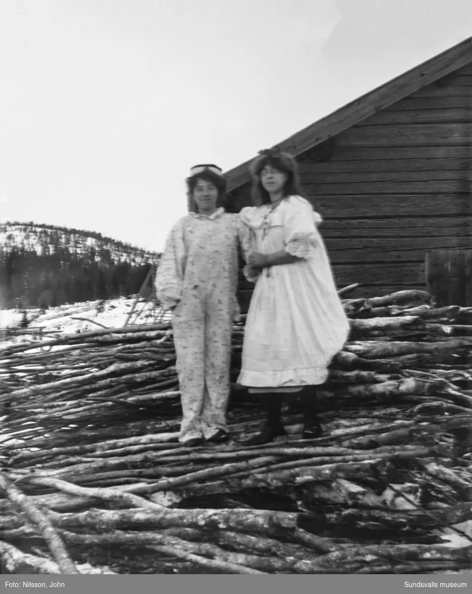 Tre bilder med två unga kvinnor som ser ut att vara utklädda och spexa lite i vinterlandskapet. På första bilden har båda löst sittande overaller, på andra bilden står de på en vedhög, den ena har nu en vit särk. På sista bilden står de på varsin sida om två andra kvinnor och en man i dörröppningen till ett timrat hus. En av dem har nu en kappa på sig.