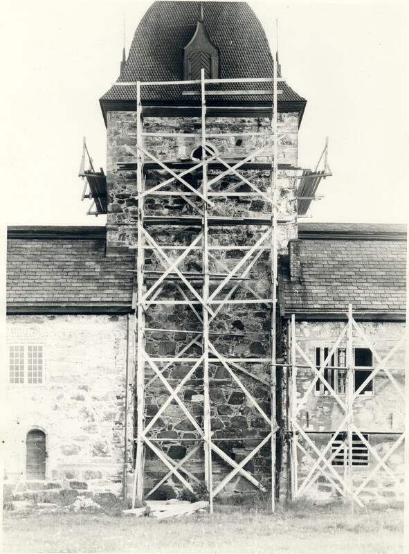 Svart-hvitt bilde med utsnitt av Austråttborgens fasade. Tårnet er midtstilt i bildet. Langs veggen, og oppover tårnet, er det satt opp et stillas av tre.