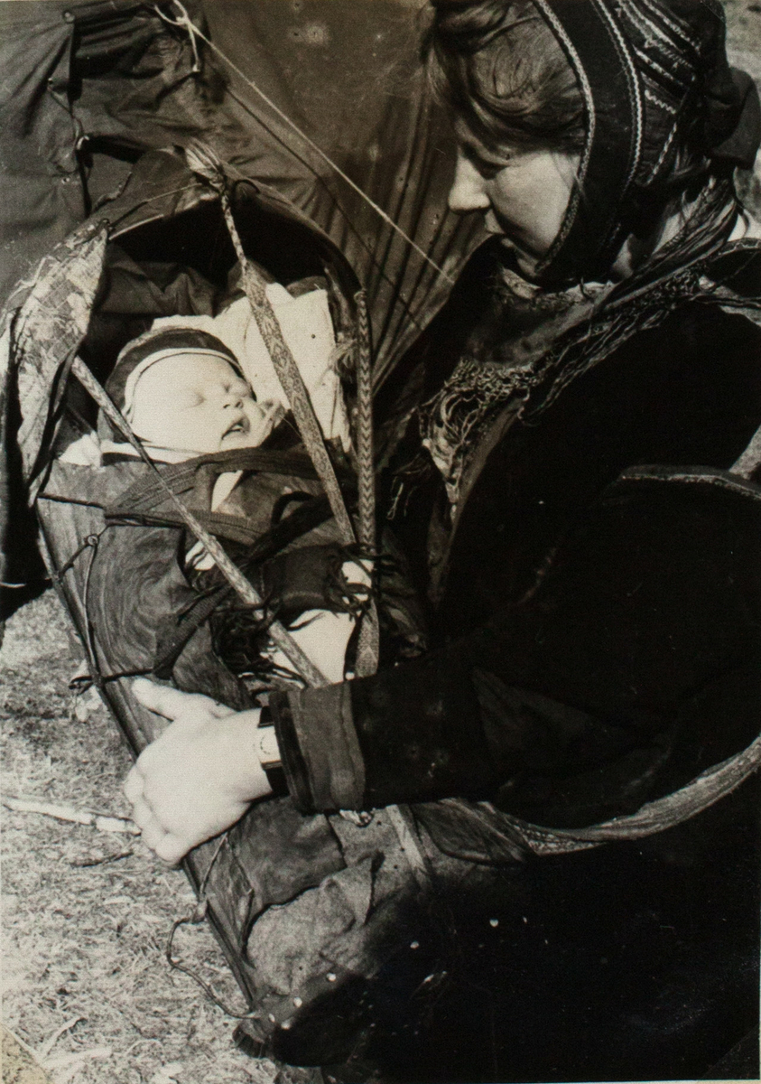 Bildet viser en samisk kvinne som holder en komse med spedbarnet inn i. Babyen sover men hans mor ser ned på den. Kvinne sitter ved siden av et telt og er ikledd tradisjonelle samiske antrekk.