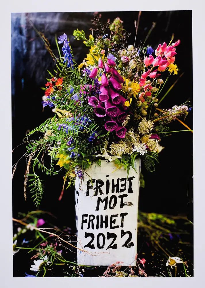 Colorful bouquet of wildflowers in a vase that is a white painted milk carton. On the cardboard is written in black letters: Freedom against freedom 2022. Photograph.