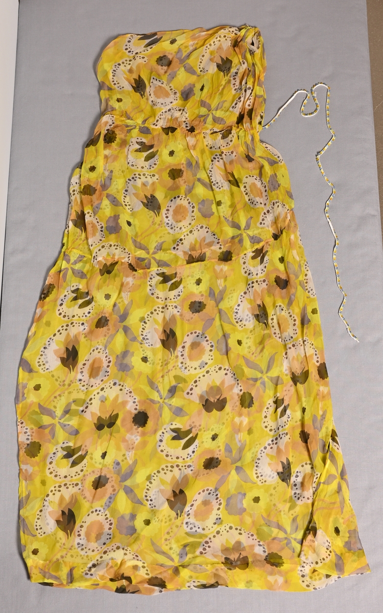 Blommig klänning av sidenchiffong, tryckt mönster i gulgrönt, brunlila, svart och brunt. Rak modell som lämnar vänster axel bar, hög midja. "Skärp" av resårband med fastsydda glaspärlor i ljusblått, ljusgrönt och gulbrunt (BM54231).