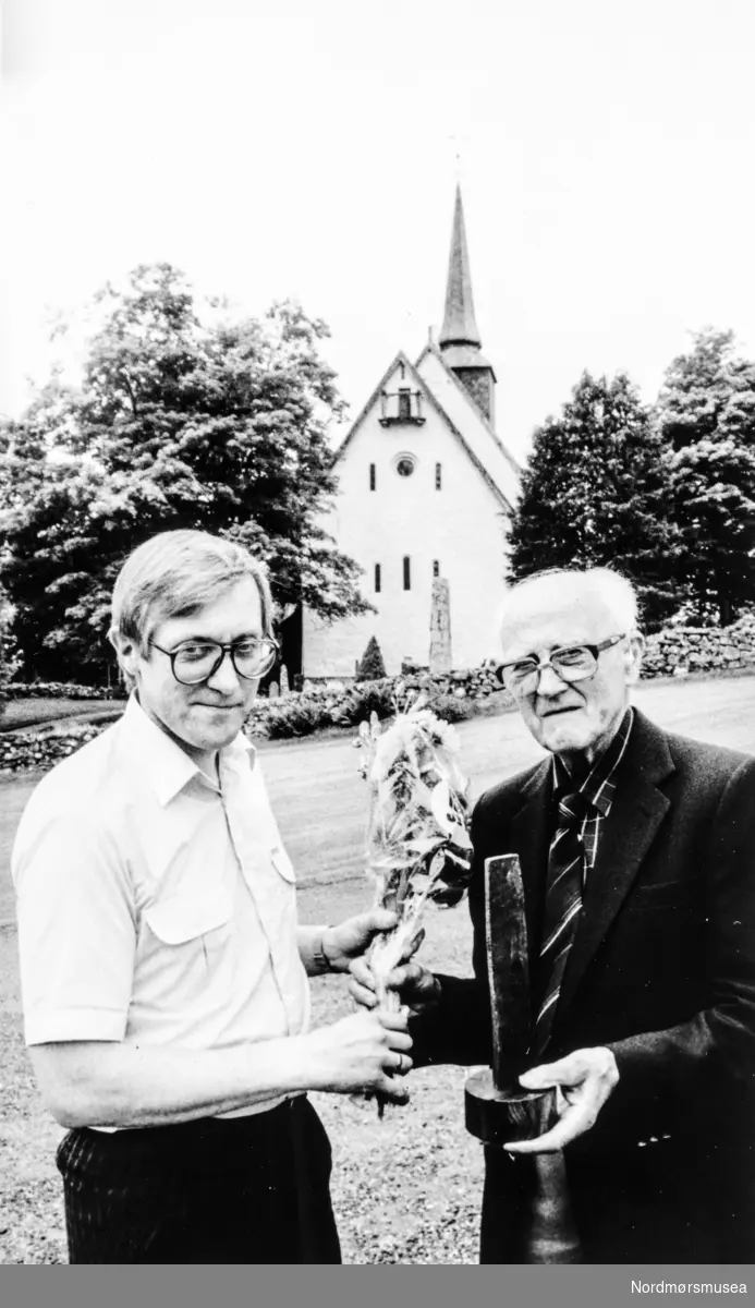Ordfører Einar Lund (til venstre) overrakte blomster til Kulipris-vinner Martin Lamvik (91 år), gammelklokkeren i Tingvoll kirke. Bildet er fra Tidens Krav, 18. juni 1988. Bildet er fra avisa Tidens Krav sitt arkiv i tidsrommet 1970-1994. Nå i Nordmøre museums fotosamling.