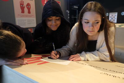 Tre jenter står sammen ved en utstillings modul. Den ene skriver på et ark.