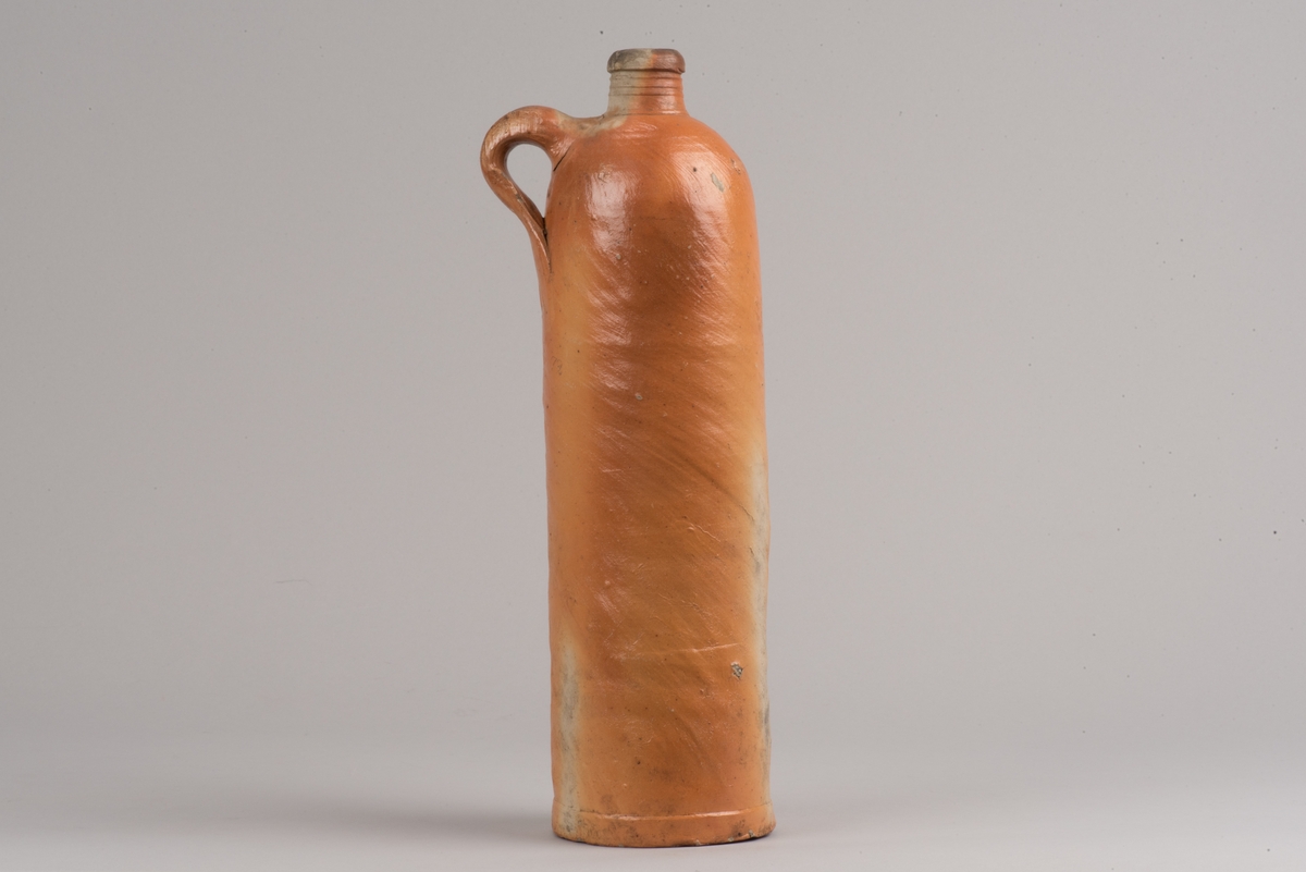 Krus i form av flaska tillverkat av saltglaserat stengods.
Cylindrisk flaska med kort hals och handtag. På buken finns två stämplar som visar att det innehållit seltersvatten.