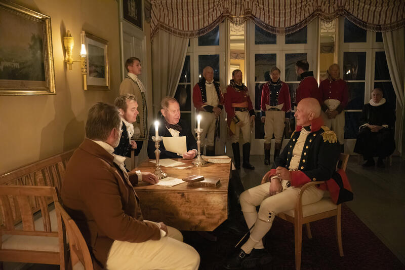 En gruppe menn i 1800-talls klær sitter og står i Hagestuen i Eidsvollsbygningen, det er kveld og rommet er kun opplyst av stearinlys og oljelamper.