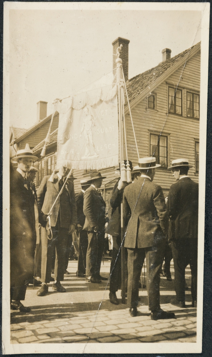 En gruppe menn i dress og hatt står rundt et banner med idrettens motto "En sund sjæl et sundt legeme". På toppen er en liten statue av en løpende mann.