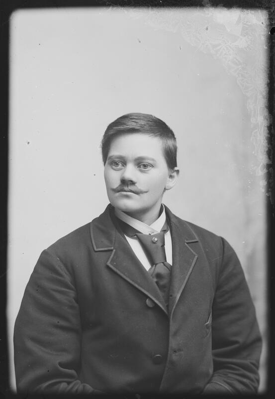 A portrait of a person with a moustache, looking slightly to the left. The person has a suit, tie, water-combed hair and brat. The person appears as a man, but is the woman Marie Høeg. Black and white photograph.
