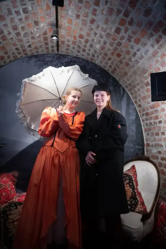 Two girls in dress-up clothes, one in an orange, old-fashioned dress and lace parasol, the other in a navy jacket. Both smile at the camera.