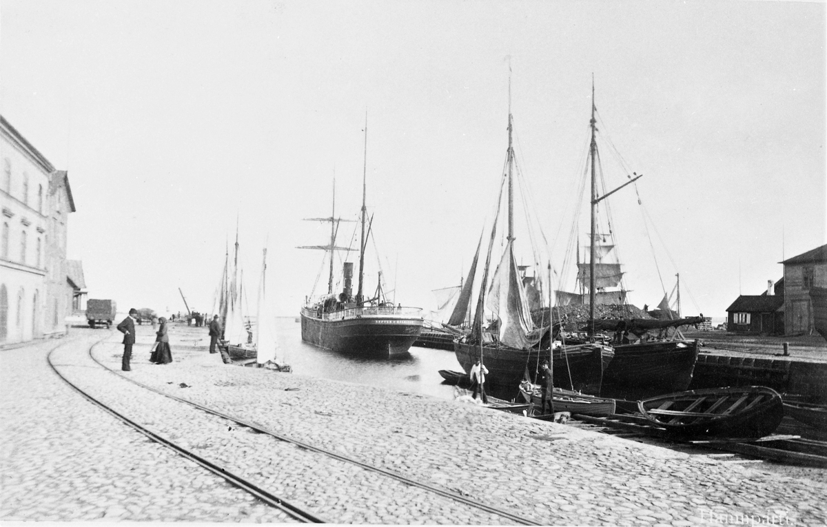 "Hamnparti" från Varberg. Hamnmagasinet till vänster uppfördes 1874. Det större fartyget längst ute i hamnen t h är ångbåten Neptun från Halmstad, som även har segel, som fraktade sten, kol, koks och styckegods. Traden var Göteborg-Lübeck. På kajen ligger räls för godsvagnarna; Järnvägen Varberg-Borås ingivdes 1880.