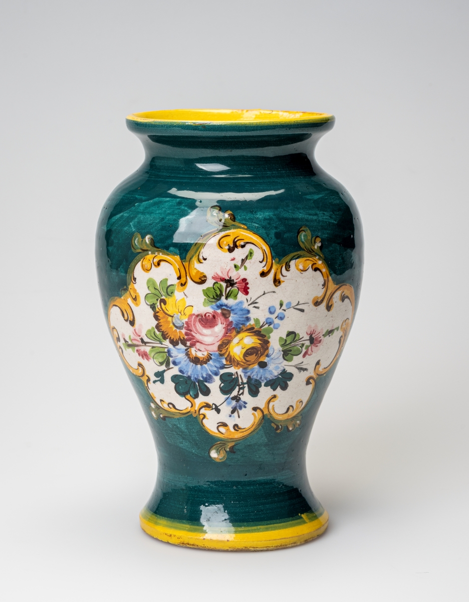 En urneformet vase av keramikk. Kanten langs foten og randen er malt i en klar gulfarge. Ellers er vasen grønnmalt. På hver side av vasen er det et blomstermotiv med blomster i gult, blått, rosa og brunt med grønne blader på hvit bakgrunn. Motivet er omkranset av enkle akantusblader i gult og grønt med detlajer i brunt. Innvendig er vasen hvit/gråhvit. På undersiden er det risset inn tekst (produsent og produksjonsland), se "Påført tekst/merker".