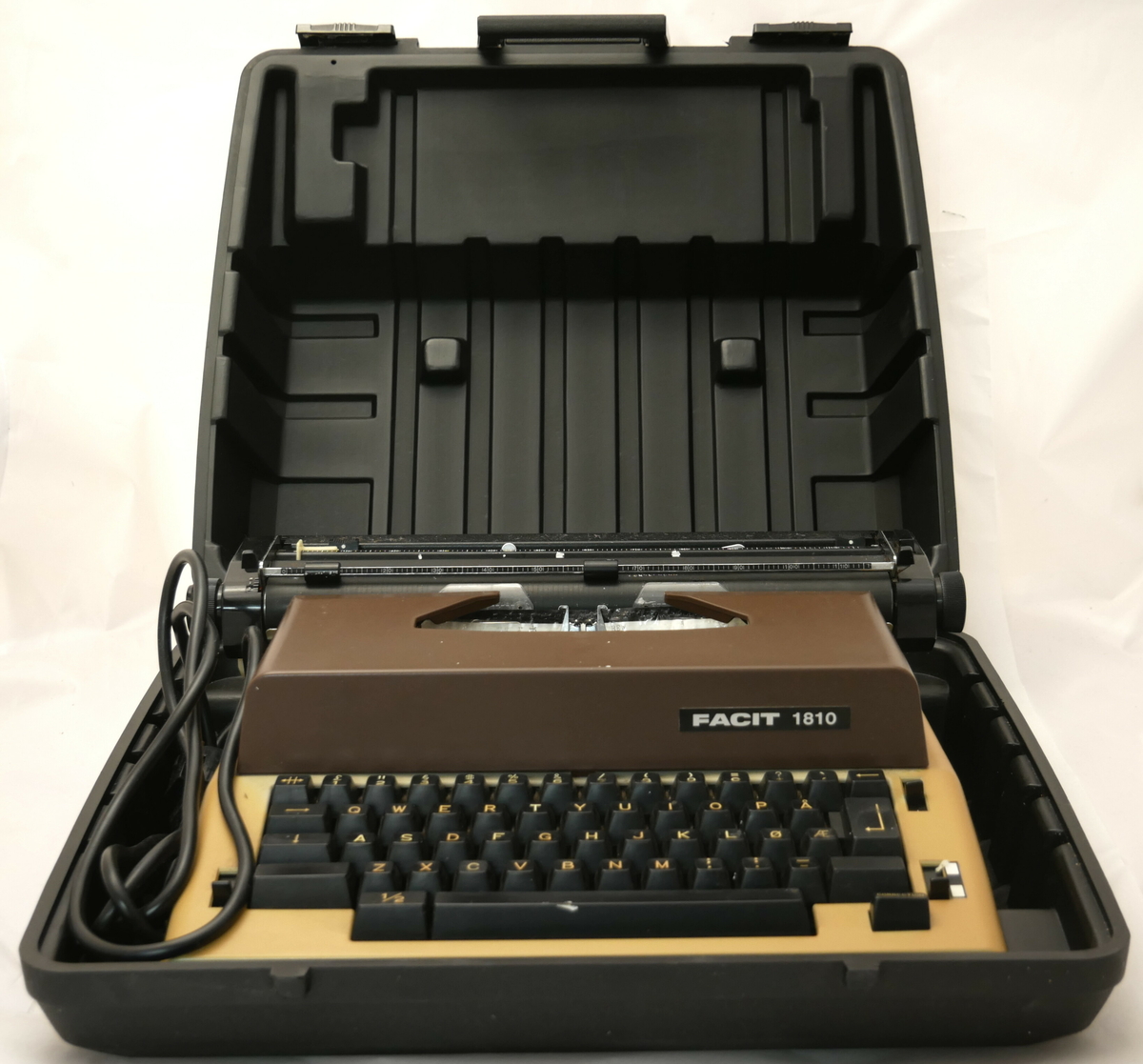 Elektrisk skrivemaskin i plastkoffert. Kofferten har handtak. Skrivemaskinen har leidning med støpsel og den kan løftast ut av kofferten.