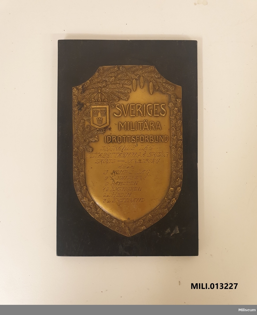 Prissköld av bronserad mässing, uppfäst på träplatta. "Segrande lag i stafettåkning å skidor, Skövde-Karlsborg 1924. 3 komp:s lag 7 Ljungberg, 9 Ramsten, 12 Ekengren, 22 Hedin, 32 Ekstrand."