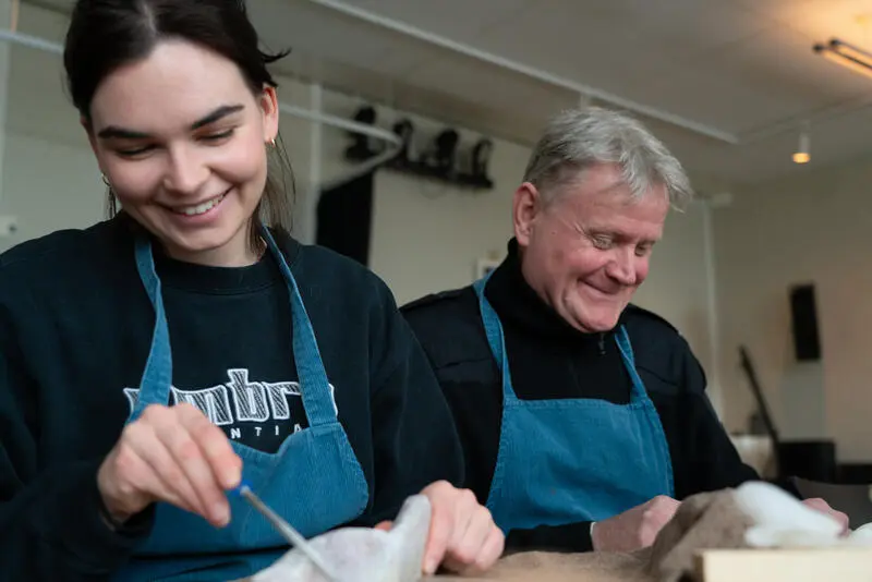 To personer, en dame og en mann sitter ved et bord og jobber med håndarbeid. De smiler