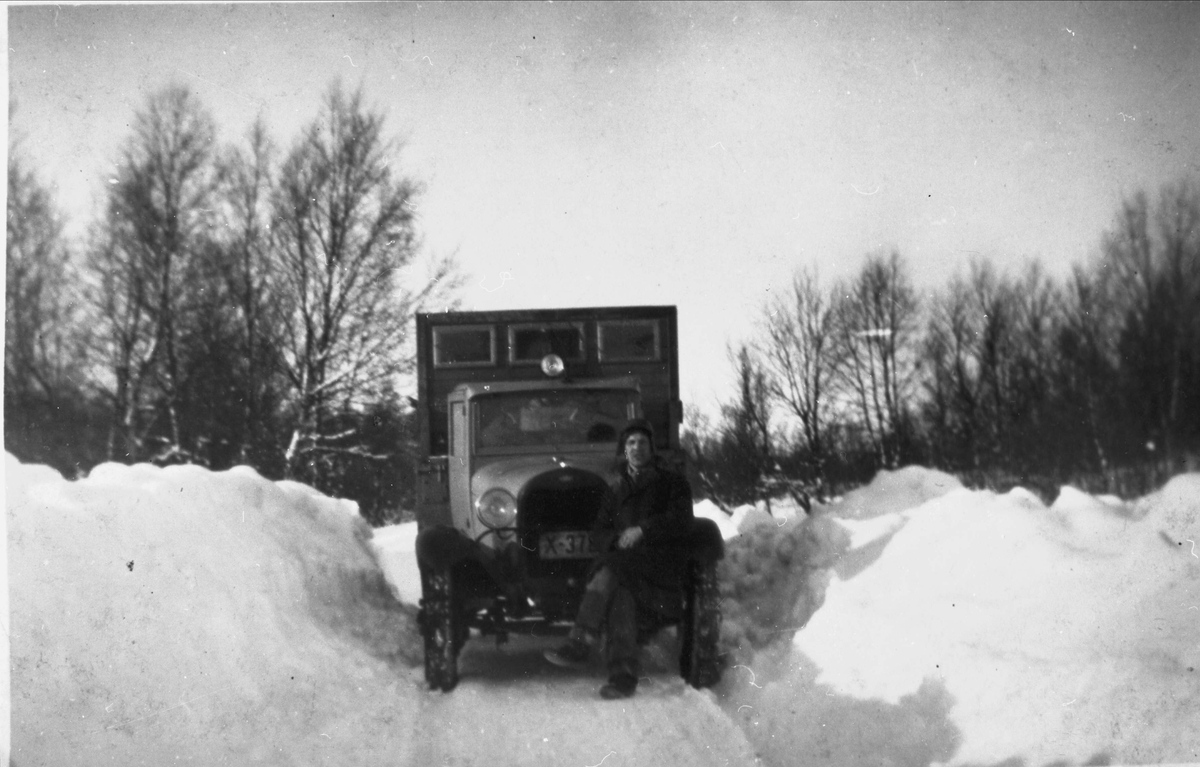 Ford lastebil på smal vintervei med høye brøytekanter. Mann sitter på støtfangeren foran.