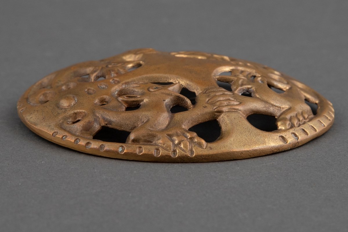 Sirkulær, lett konveks brosje i bronse med symmetrisk, gjennombrutt ornamentikk med fire fabeldyr. Nål på baksiden.