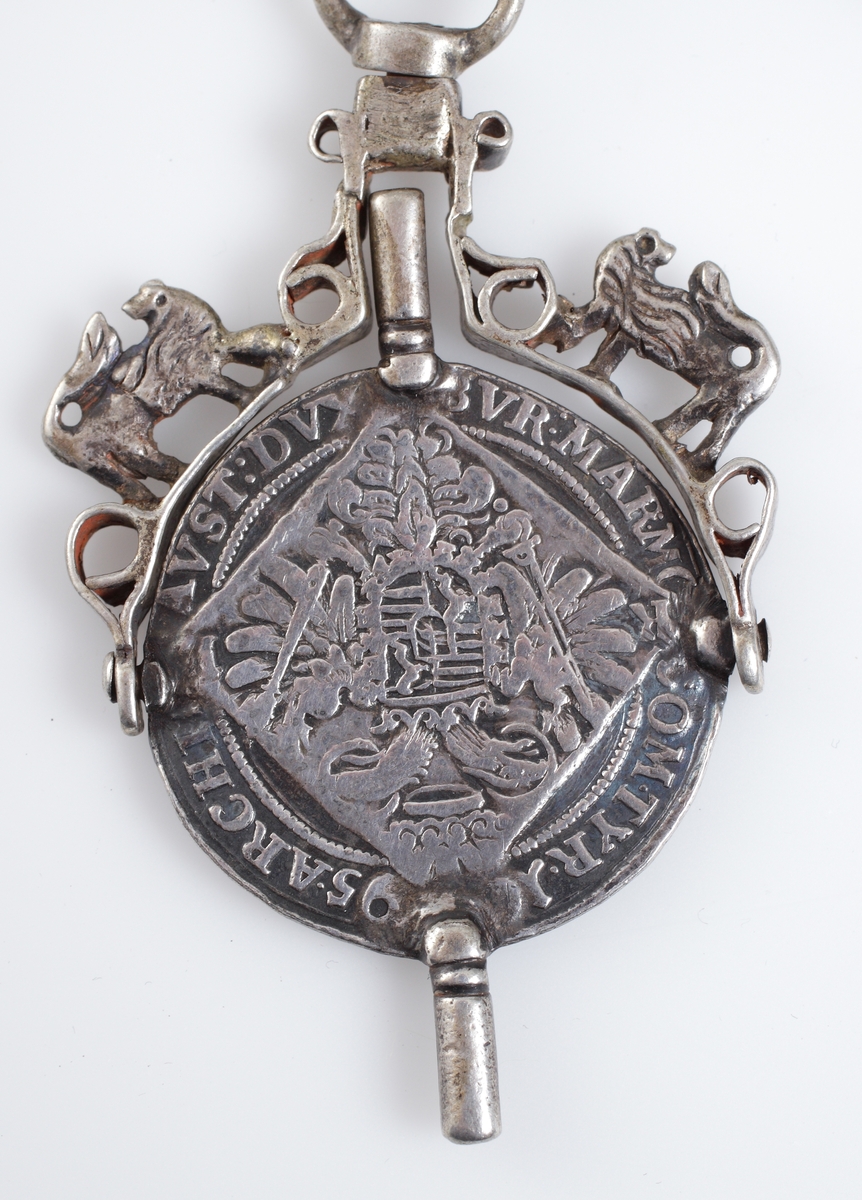 Chatelaine av silver. Kortare dekorerad kedja försedd med två urnycklar, signet (A.E.), amulett i filigranarbete och elfenben, vari antagligen suttit en bit av korall.

Äldre, österrikisk tillverkning.

Inskrivet i huvudkatalog 1909.