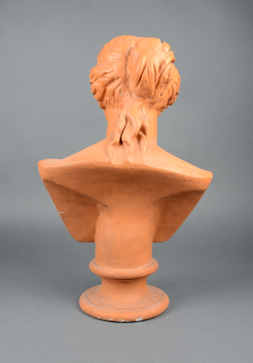Byste av Venus med klassisk ansikt og frisyre. Den står på rund sokkel og er terracottafarget.
