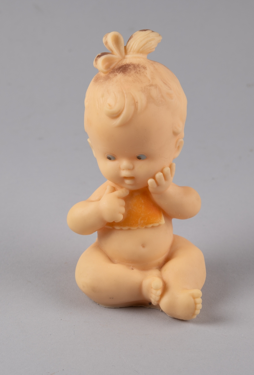Leketøy av mykplast i form av en sittende baby. Den har orange bleie og smekke, blåe øyer og håret er satt opp i en sløyfe.