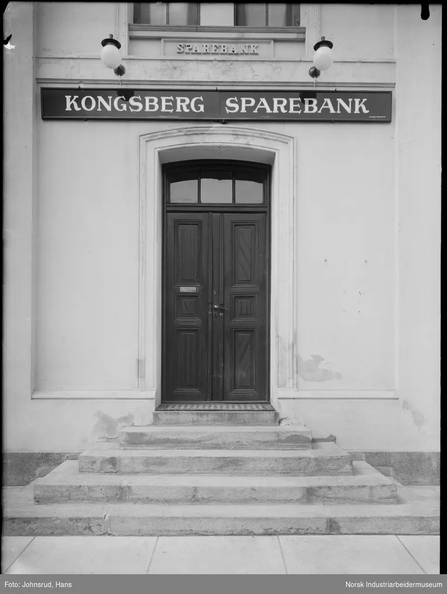 Inngang til Kongsberg Sparebank. Trapp i mur opp til dobbel dør i tre. Skilt og belysning over døren.