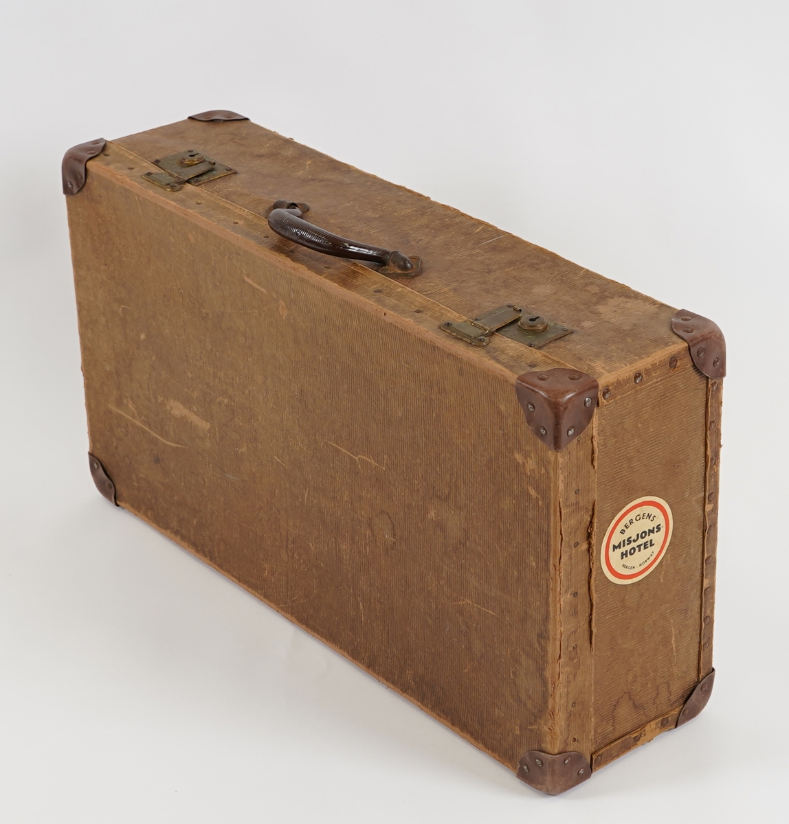 Ein rektangulær koffert, laga av pappliknande materiale, på ei innvendig treramme. Hjørna er forsterka med skinn. Handtak, henglser og låsar er i metall.