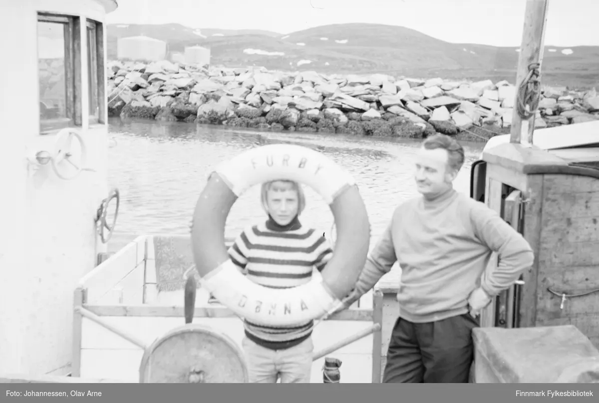 Furøy av Dønna ring på havna i Båtsfjorden, Mobil Oil i bakgrunnen

En gutt holder på en livbøyle med skriften "Furøy Dønna" og mann står ved siden av 