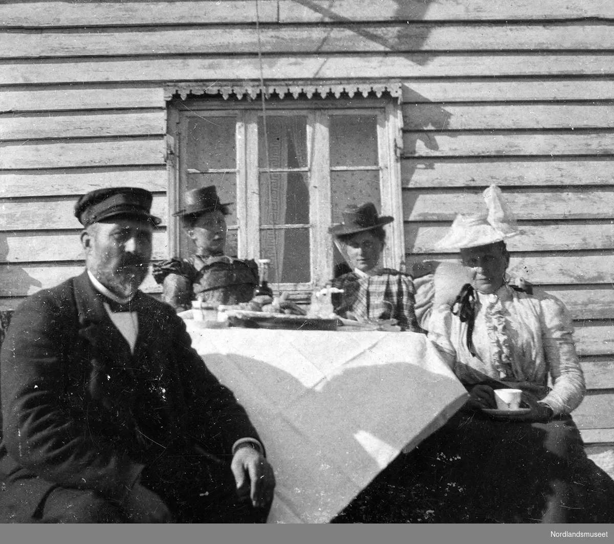 En mann og tre damer samlet rundt et kaffebord ute ved veggen til et hus på gården Fjellbirkeland, Bergen. Et vindu bak dem.