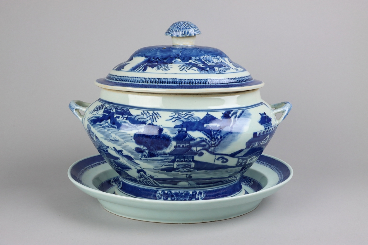 Oval shaped lid with slightly domed form, on top a knob in shape of a flower head. Around the knob lotus leaves, surrounded by a list of pagoda landscapes. The edge of the lid with dark blue border in a criss cross pattern. Oval lobed bowl with angular handles, supported on a central fot. On both sides of the bowl decor of landscape scenes of pagodas, buildings, gardens and waters. Handles in shape of bent tree branches. On the foot a dark blue border with rectangular reserves filled with symbols of good fortune. Oval stand with a lip decorated with dark blue border with criss cross patterns and chrystanthemum flowers. The everted rim is decorated with a dark blue border with a similar pattern as above, but complemented  with oval reserves filled with symbols of fortunes. The well is decorated with landscapes scenes of pagodas, buildings, gardens and bridges. All decor in blue underglaze.