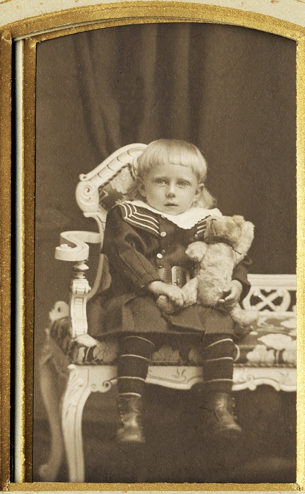En pojke i sjömansdräkt sitter på en liten vitmålad soffa med en nalle i famnen.
Helfigur, en face. Ateljéfoto.