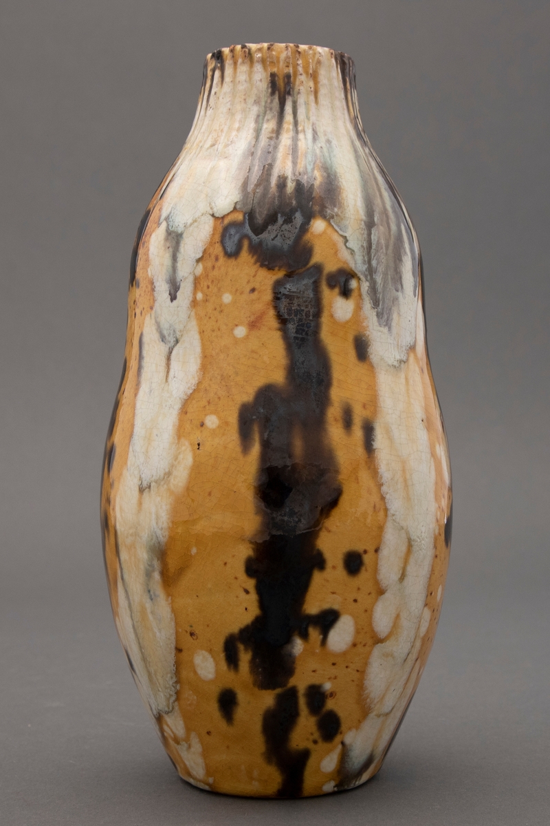 Dreid vase med lett antydet kalebassform. Rifler ned fra munningskanten. Gulglasert grunn brune spetter. Svart og hvit rennedne glasur nedover vasen.