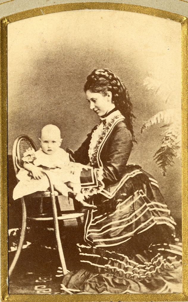 En ung kvinna i klänning med turnyr, tillsammans med sin son.
Helkropp. Ateljéfoto.