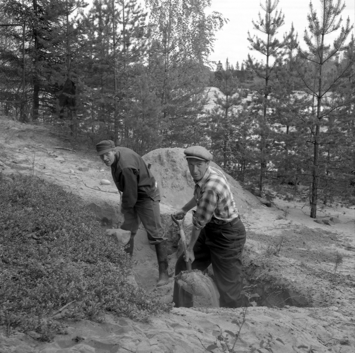Bygging av hengebrua over Prestfossen mellom Glomdalsmuseet og Prestøya.
Gravearbeid, to menn.
Det kostet kr.120000 for å bygge 130 meter lang hengebrua i 1958.