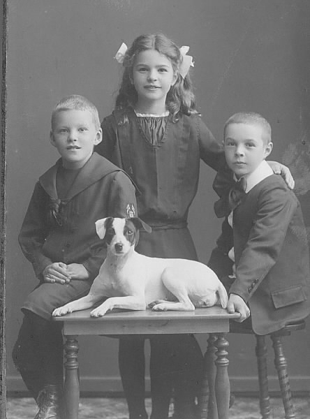 Ateljéfoto av barnen till handelsman Oscar Pehrson; en flicka och två pojkar. Framför barnen ligger en liten hund på ett bord.