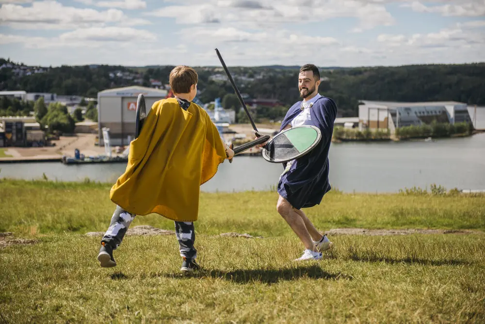 En ung gutt og en voksen mann kriger med sverd og skjold på toppen av et fjell. Gutten har gul kappe på seg og står med ryggen mot kameraet.