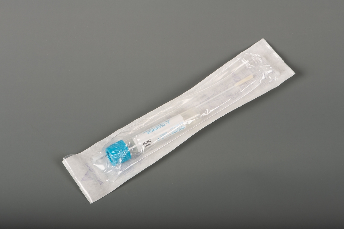 En plastlomme limt på et papir inneholdende en tube og en pinsett. Ten inneholder en klar væske, har blå kork og en etikett med skrift i svart og blått.