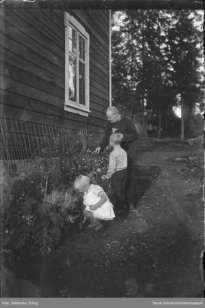 Mann i Frelsesarmeen uniform plukker blomster sammen med to barn utenfor bolighus.