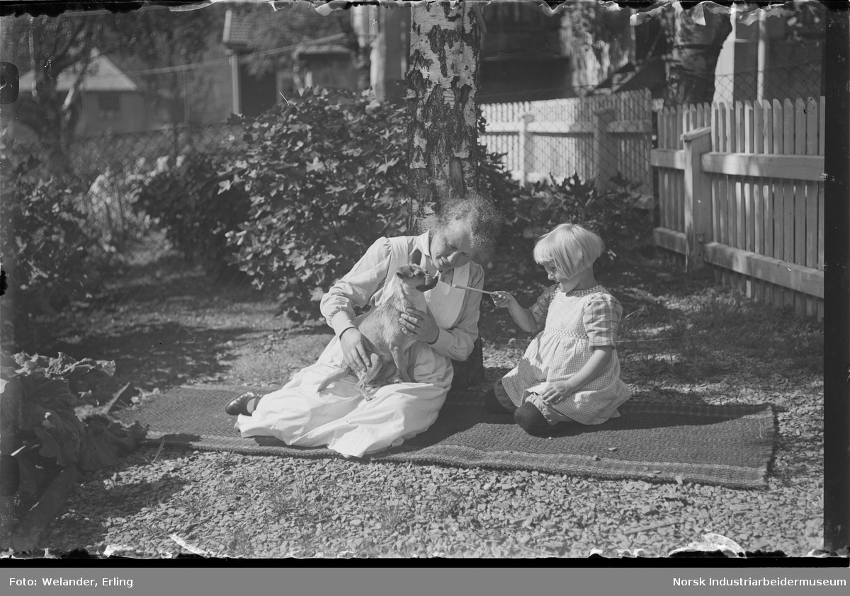 Kvinne og barn sittende på teppe i hage. Kvinnen har en hund på fanget som barnet gir noe til. Elevheimen i bakgrunnen.