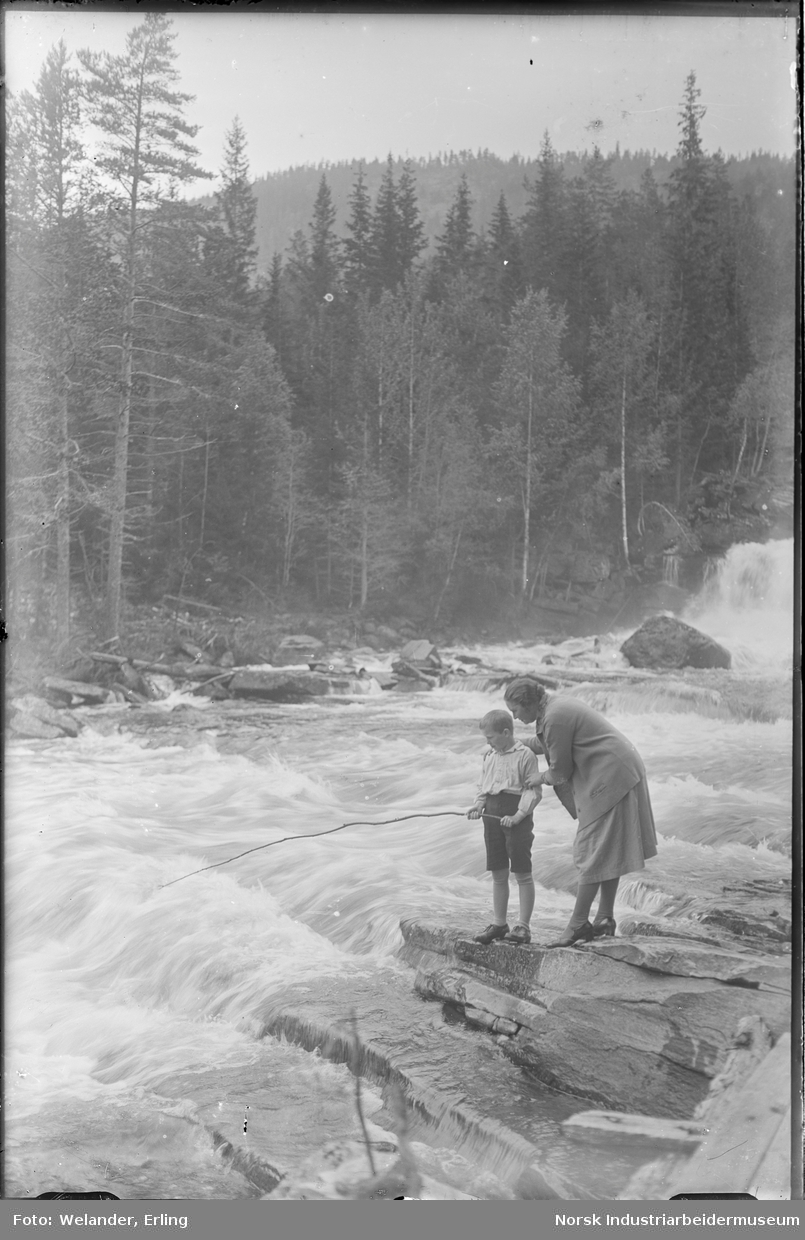 Kvinne og barn stående ved elvestryk. Gutten holder en hjemmelaget fiskestang i hendene. Kvinnen er kledd i kjole og høye hæler, gutten i shorts og skjorte.
