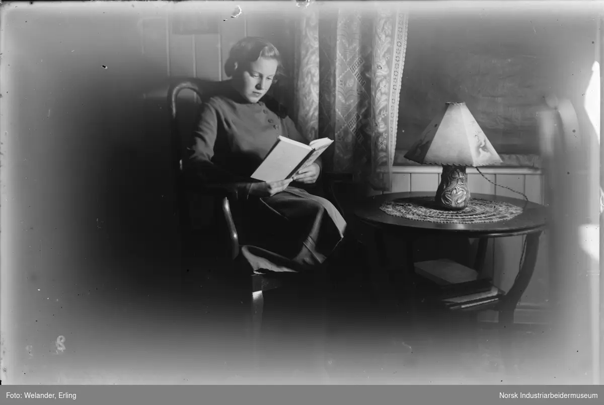 Kvinne sittende i stue og lese bok. Heklede gardiner og blendingsgardin foran vinduet. Rundt bord med lampe på foran vinduet.