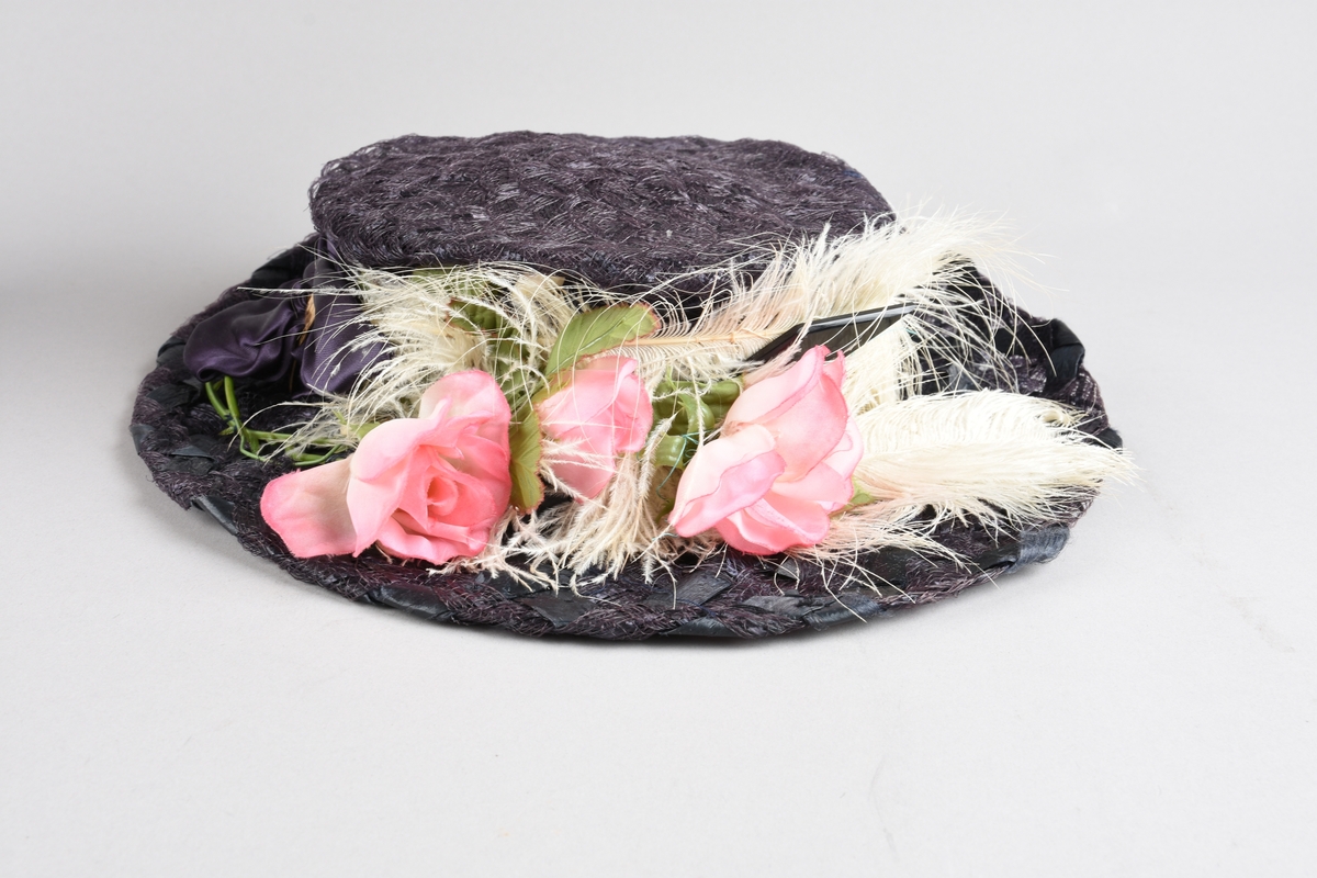 Mørkfiolett, flatpullet og bredbremmet hatt i  "bretonne-" el. "canotier-"fasong. Laget av nettvevede, kunstige stråbånd. Rundt pullen er et "hattebånd" av bred drapert sateng i samme farge, festet med spenne av hamret messingblikk. Senere laget om til kanutt-kostyme, med kunstig rosegren oppå offwhite strutsefjær dandert over pannen. Inni pullen er en skulderpute satt fast med sikkerhetsnål.  Hårnål og hatte-elastikk til feste av hatten.
