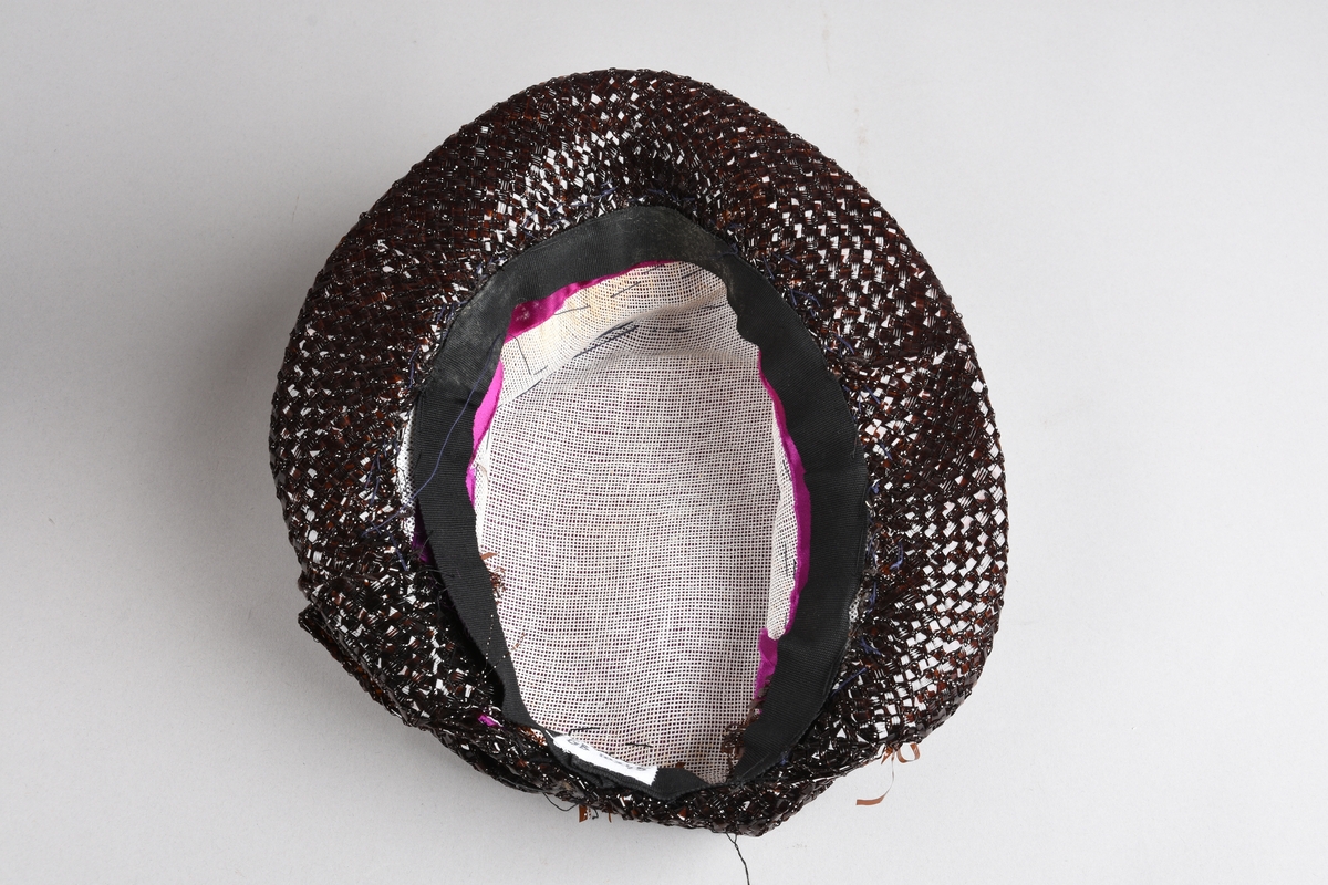 En purpurfarget hatt av flat satengpull med folde-drapert kant, omspennes av en lav, kobbersort, flettet cellofan-"karm" og smal brem. I nakken er det en bred, flat sløyfe av cellofanstoffet. Bremmen er senere avskåret i nakken og sydd opp. Over pannen viser trådender at noe pynt er tatt av (rosene og strutsefjæren på den senere hatt GB 18344?). Inni er hatten avstivet med stramei-liknende stivstoff og er isydd et hattebånd.