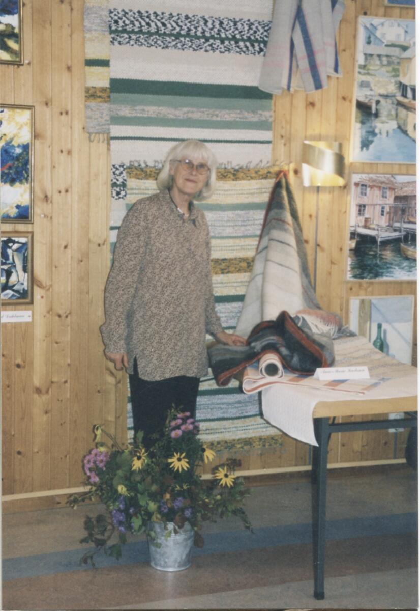 Hantverksutställning i Missionskyrkan cirka 1980. Ann-Marie Karlsson (1929 - 2020) står vid vävt material. Bakom henne på väggen hänger vävda mattor.