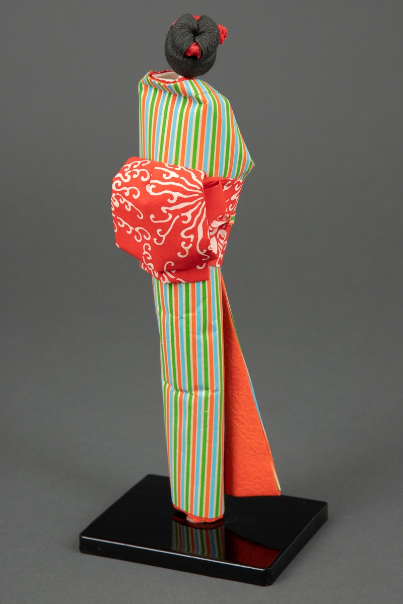 Håndlaget papirdukke i japansk bretteteknikk plassert på en svartmalt tresokkel. Alle deler er laget i papir bortsett fra "kroppen" som består av en trepinne og vatt. Dukken har rød blomst og hårpynt i oppsatt hår og er iført en kimono med vertikale striper i hvitt, oransje, blått og grønt. Rød innerkrage med hvite blomster og oransje innerkimono. Dukken har på seg rød obi med stilisert skymønster og obisnor i samme mønster som kimonoen.