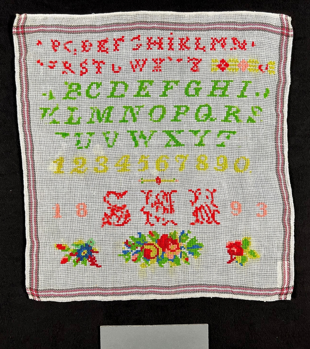 Märkduk på vit botten med korssömsbroderi i rött och grönt. Alfabetet och siffror broderade i rader, Undertill ett monogram: S.M.N. och årtalet 1893. Längst ned blommor.