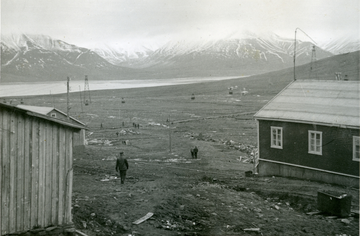 Hus i Longyearbyen i forgrunnen. Taubanen med taubanebukker/tårn til  til Gruve 2 a og 2 b i 1940/41. Det er bygd en gangbro over elva mellom byen og Sukkertoppen. Bildet er fra Jens A. Bay sin bildesamling. Han overvintret på Svalbard i 1940/41.
