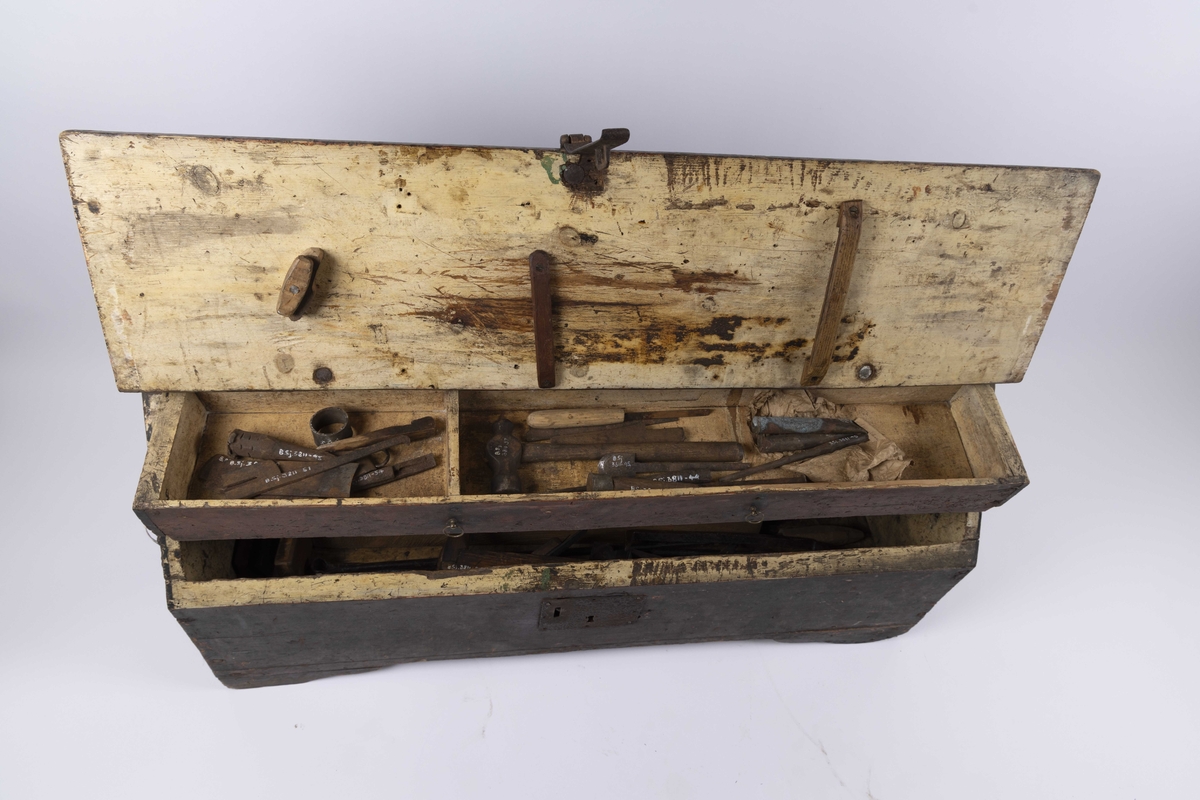 Tømmermannskiste med skuff og nøkkel. Kisten inneholder diverse verktøy, som bl.a. høvler, bor, navar, hammere, filer og stemjern. De utgjør til sammen 60 ulike verktøy.
